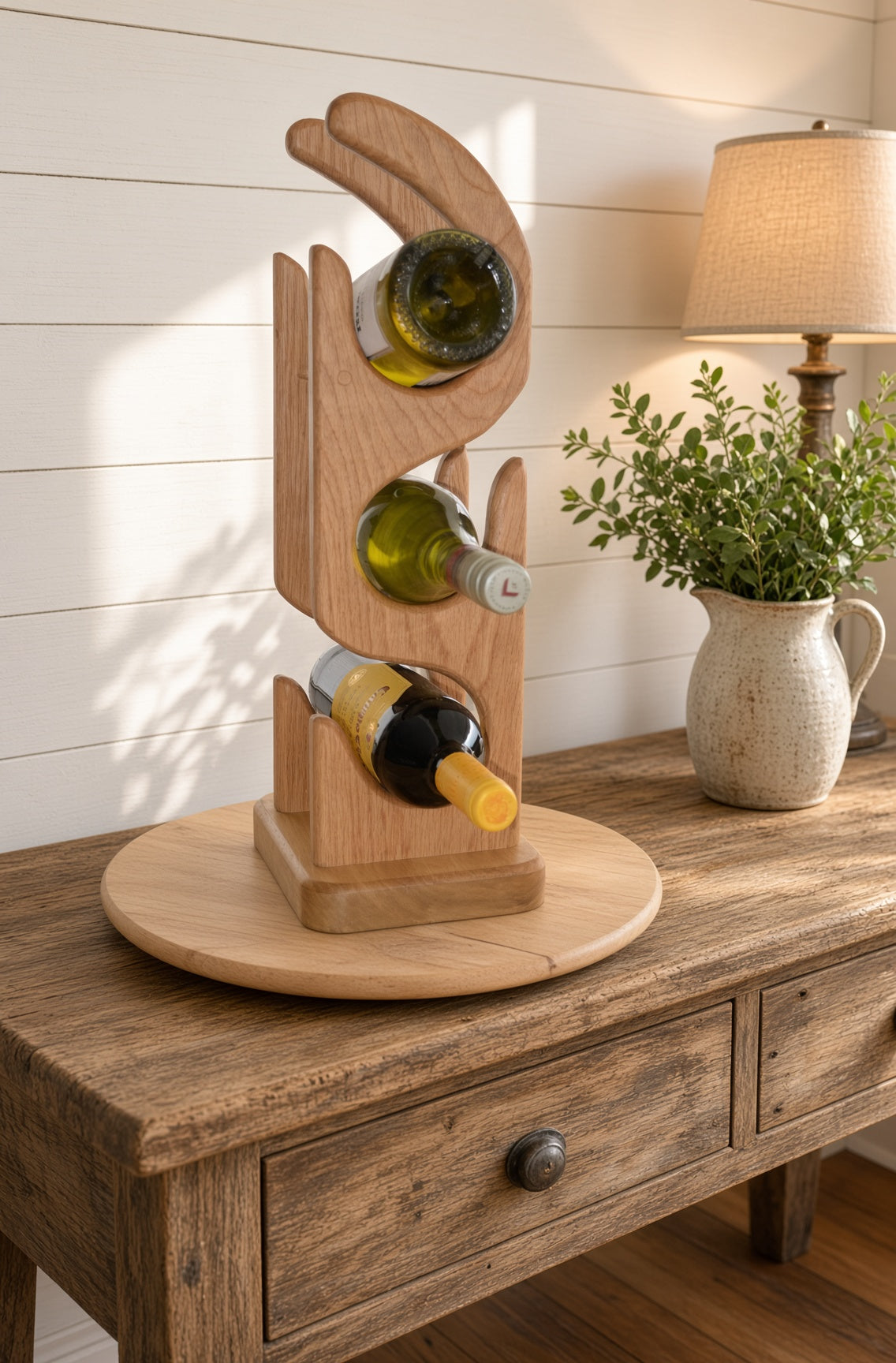 A three-tier wooden wine bottle rack shaped like a stylized hand, holding three wine bottles, displayed on a rustic wooden sideboard next to a lamp and a potted plant.